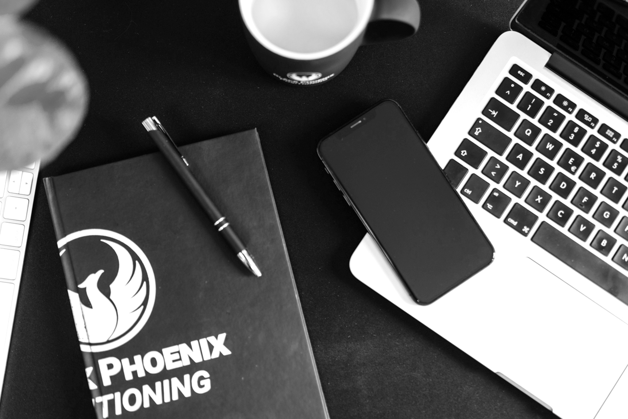 Headerbild Ein Schwarz-Weiß-Foto eines Arbeitsbereichs mit einem Laptop, einem Smartphone, einem Notizbuch mit der Aufschrift PHOENIX, einem Stift und einem Kaffeebecher auf einer schwarzen Fläche.