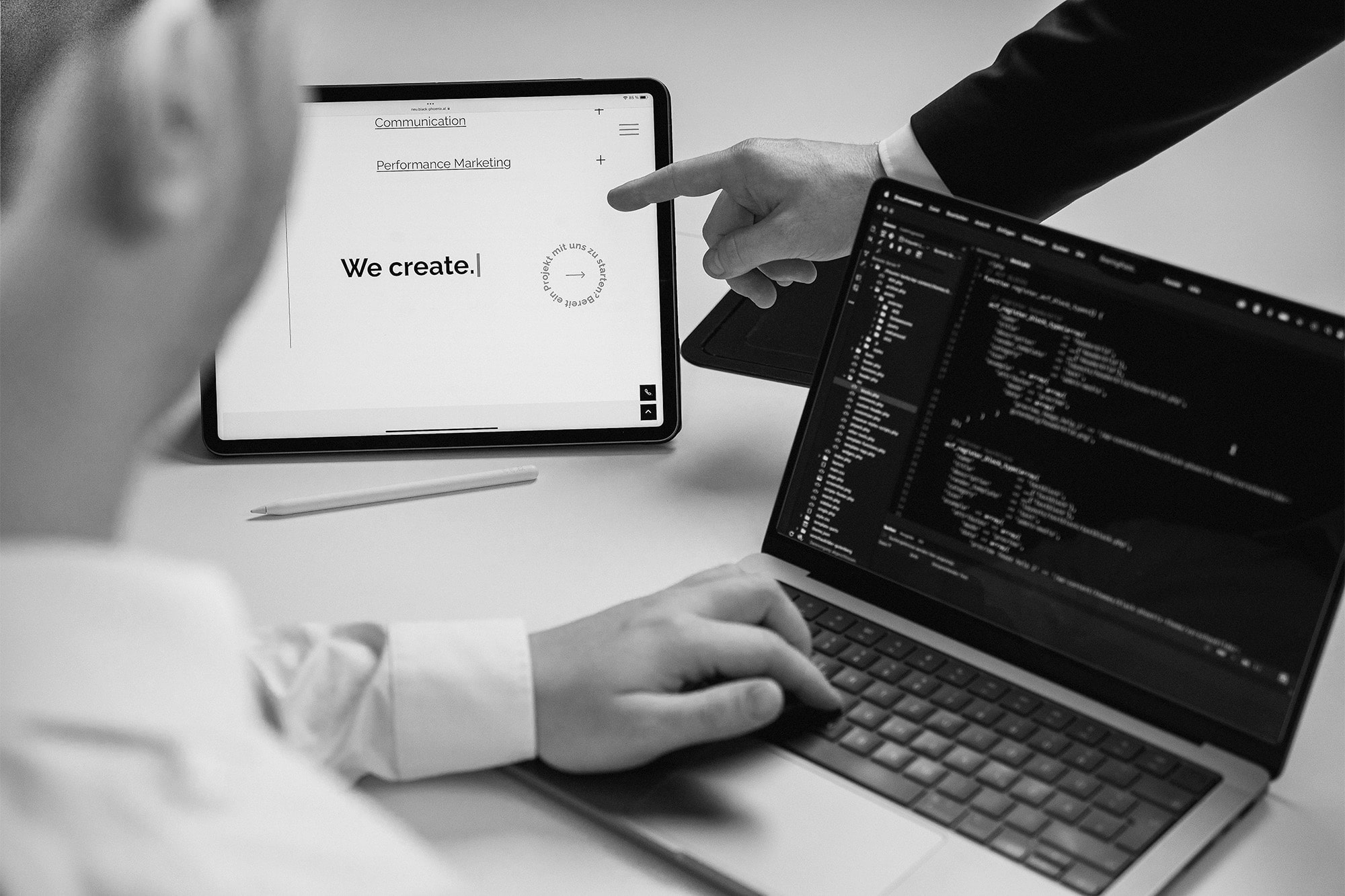 Headerbild Zwei Personen arbeiten an einem Schreibtisch; eine tippt einen Code auf einem Laptop ein, während die andere auf ein Tablet zeigt, das einen Bildschirm mit dem von uns erstellten Text und Navigationsoptionen anzeigt. Die Szene ist in schwarz-weiß gehalten.