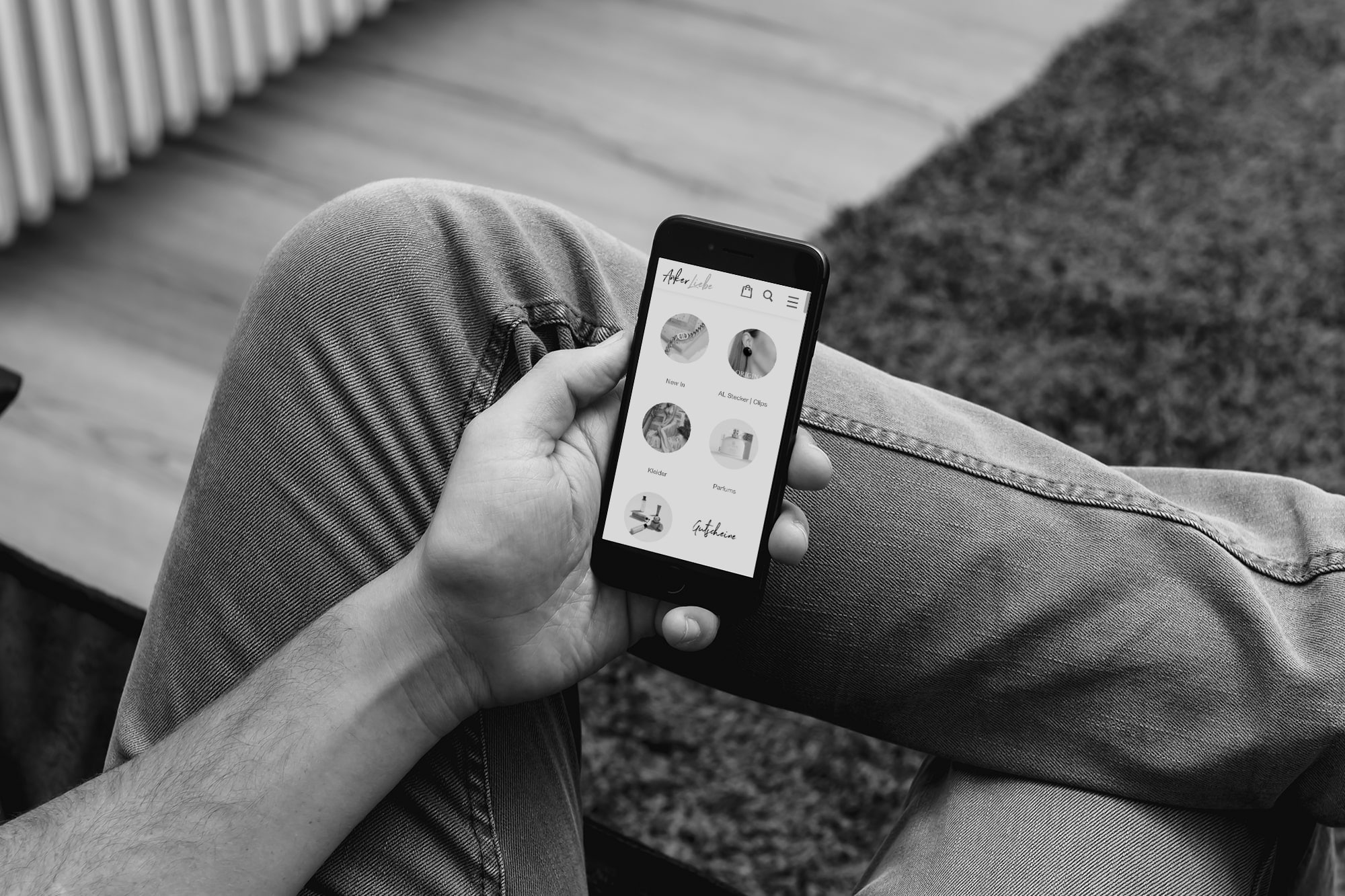 Headerbild Eine Person sitzt mit gekreuzten Beinen, hält ein Smartphone in der Hand und surft in einer Social-Media-App, die Profilsymbole und Namen anzeigt. Das Bild ist in schwarz-weiß gehalten.